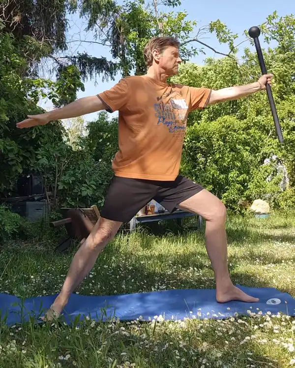 Werner Hauschild im Krieger II auf einer Yogamatte im Garten, der Steel Mace in der ausgestreckten rechten Hand