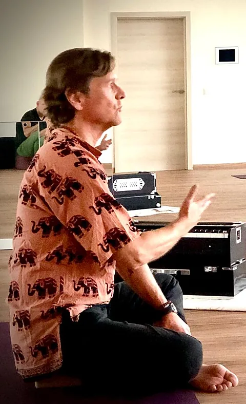 Werner Hauschild sitzt im Schneidersitz am Harmonium, eine Hand erhoben, und singt