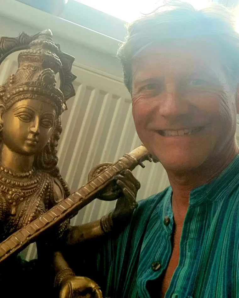 Werner Hauschild lacht in die Kamera, den Arm um eine Messingfigur der Göttin Saraswati mit Vina gelegt